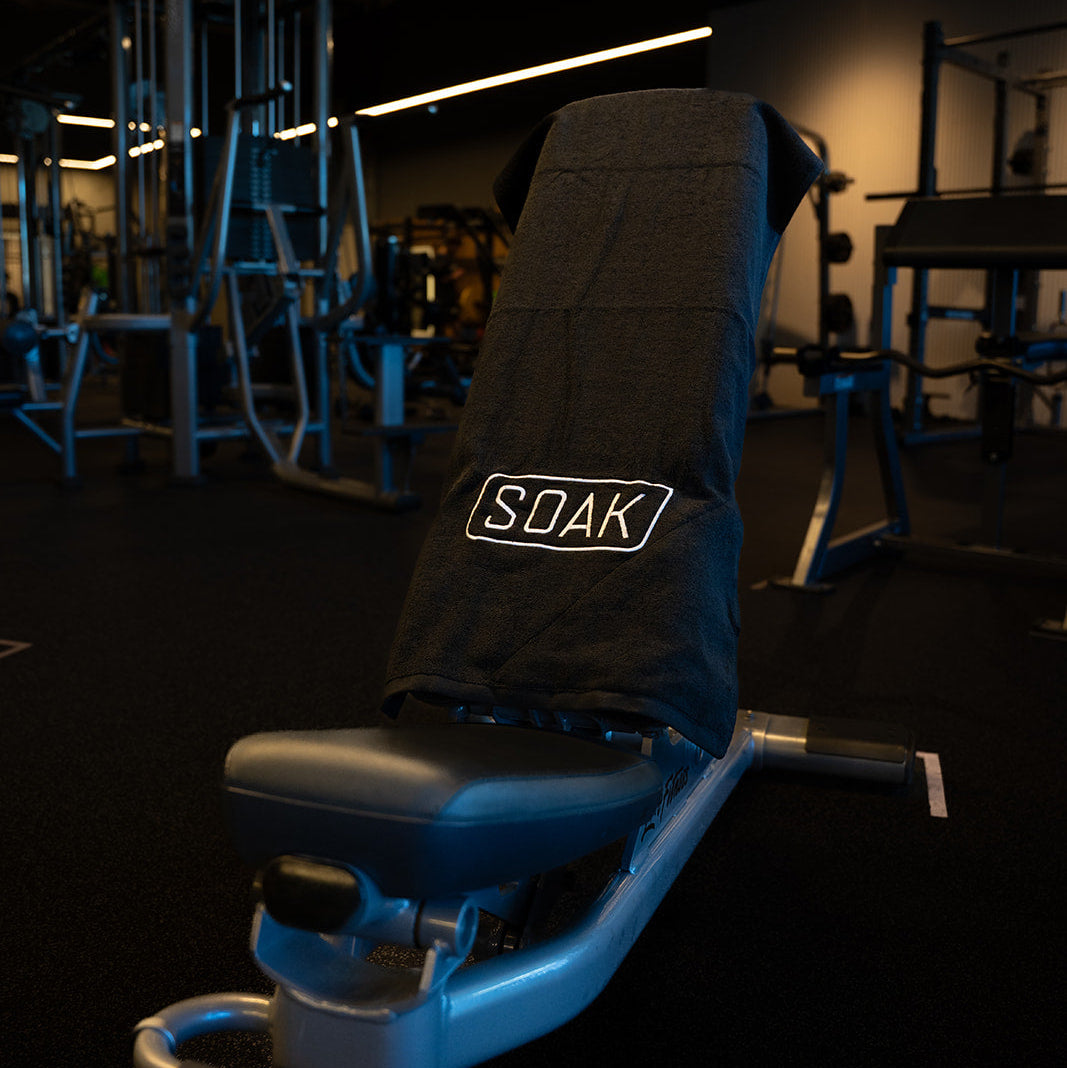 A gym weight bench with a black towel draped over the backrest, featuring the brand name 'SOAK' in white text. The background shows a dark gym interior with other exercise equipment.