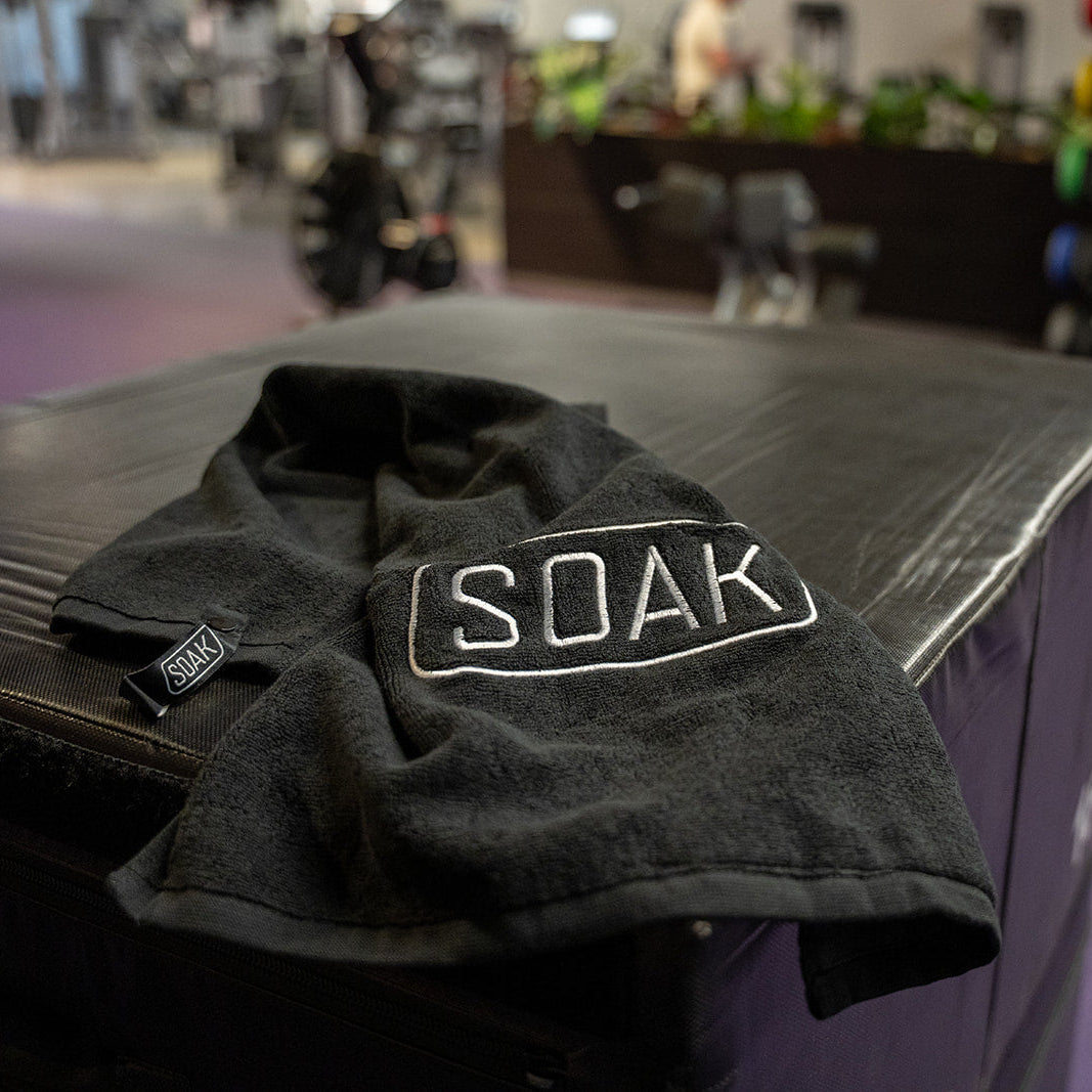 A black towel with 'SOAK' embroidered in white on the front, draped over a dark bench in a gym setting with purple flooring.