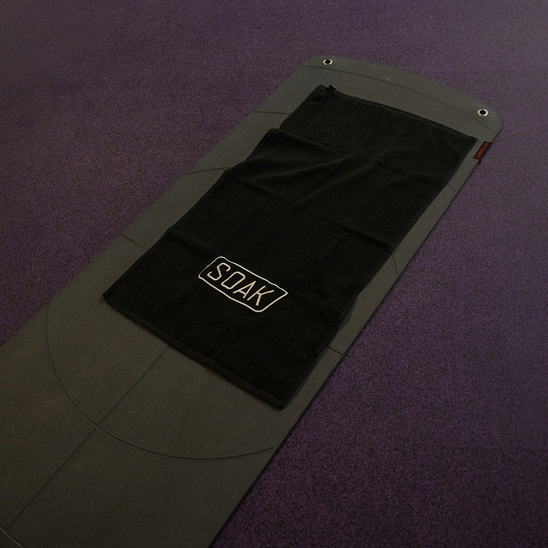 A gray and black yoga mat with a black towel featuring the 'SOAK' logo, laid out on a purple gym floor.